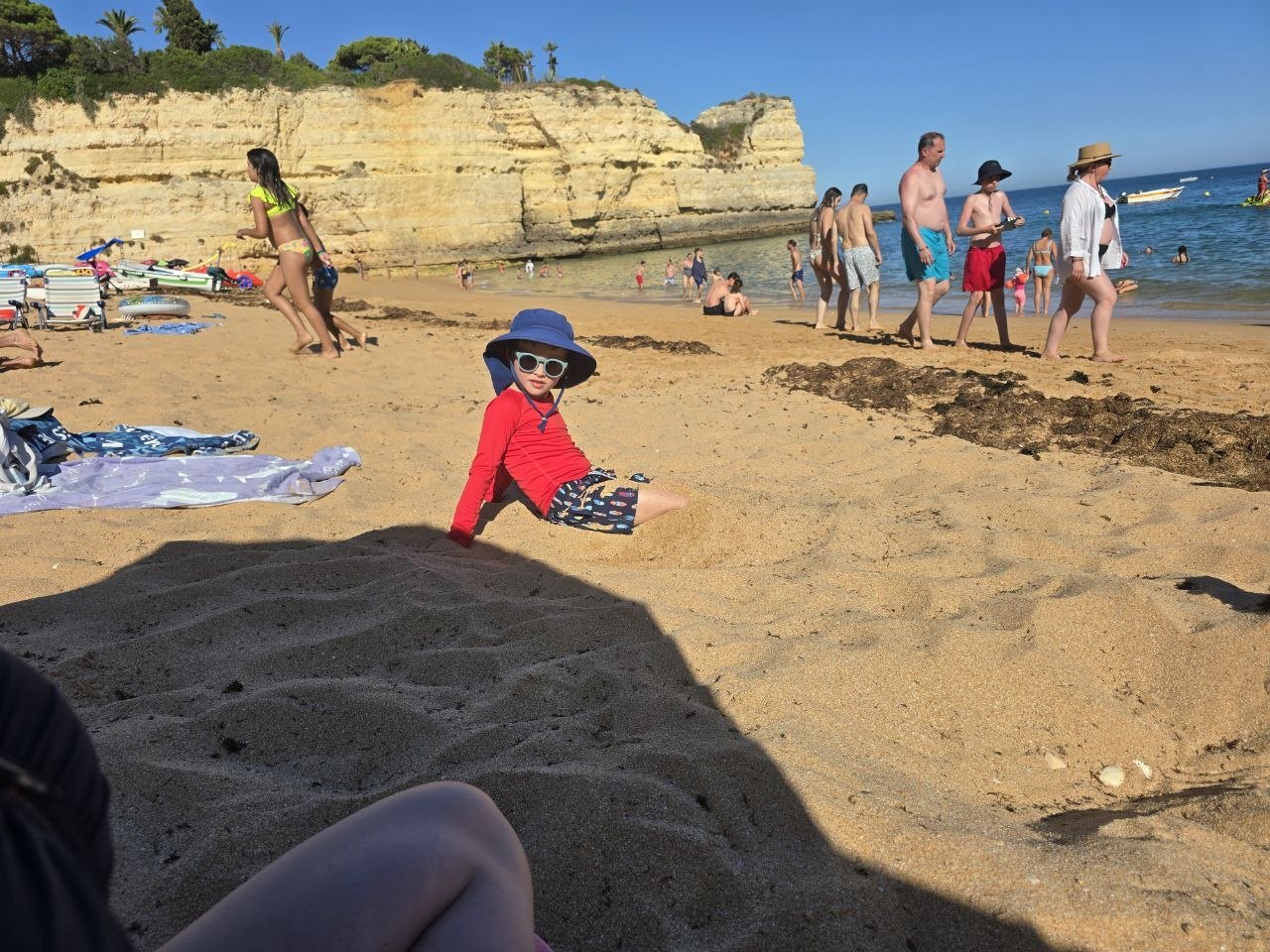 Noah sitting in the sand at Praia de Nossa Senhora da Rocha wearing a red swim shirt blue hat and sunglasses