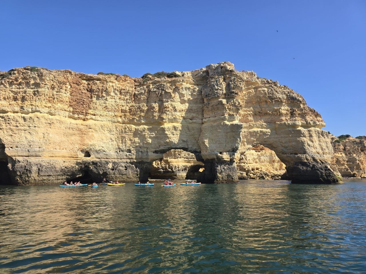 Kayakers paddling below tall golden Algarve cliffs reflected in calm water
