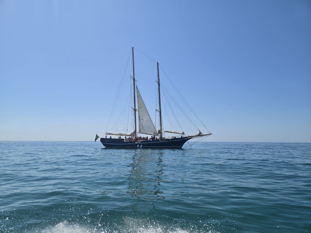 A black two-masted sailboat on calm blue water under a clear sky