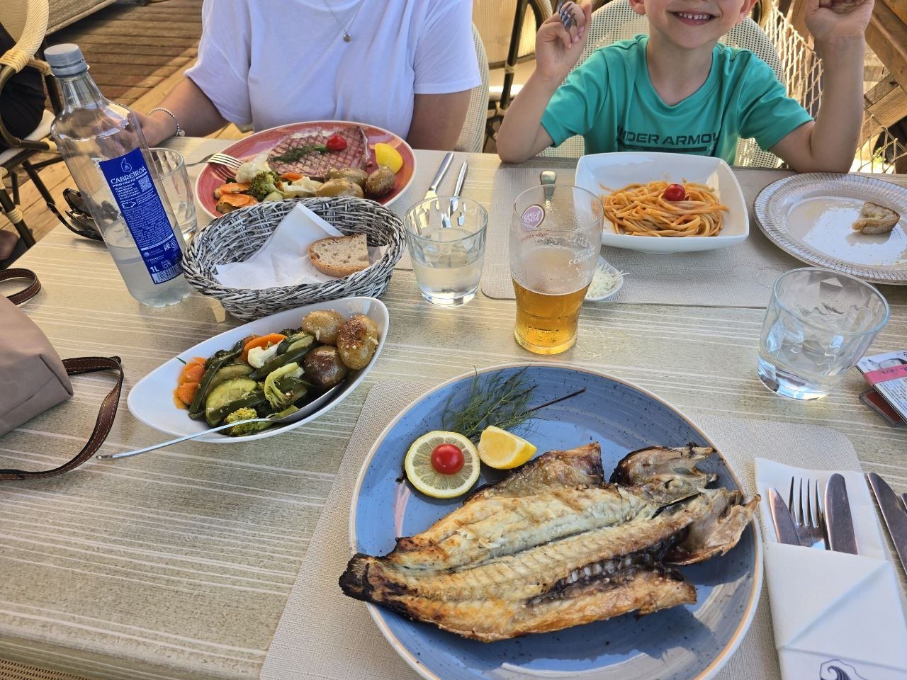 A beach restaurant table with grilled whole fish, vegetables, pasta, bread, water, and beer