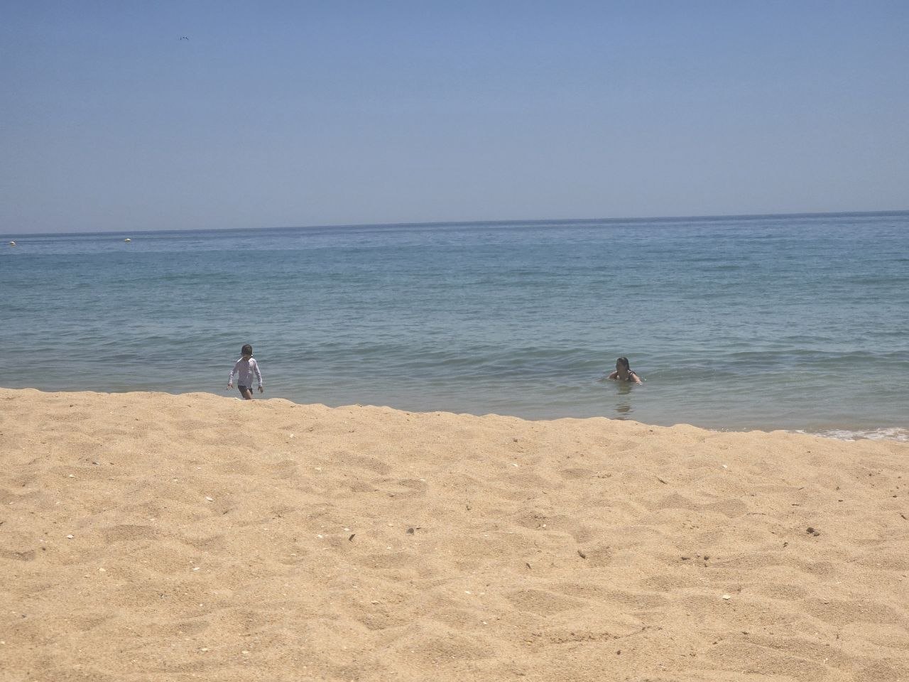 A sandy beach with Catherine and Noah in the calm blue water near shore