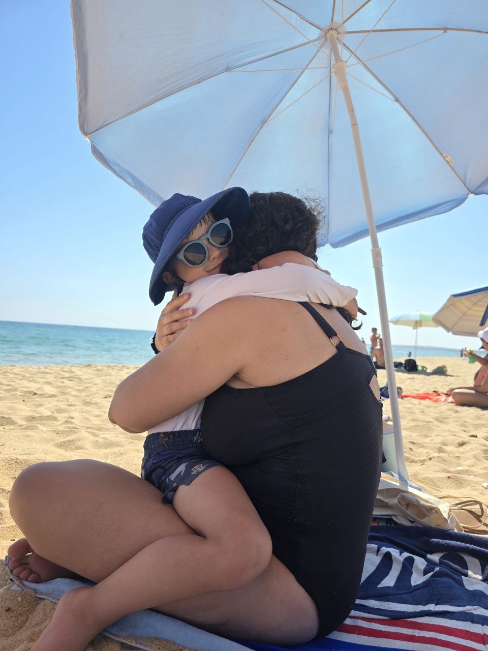 Catherine hugging Noah under a blue beach umbrella with the ocean behind them