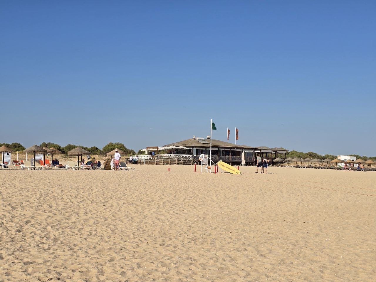 Wide sandy beach view toward a beach restaurant and straw umbrellas under a clear blue sky