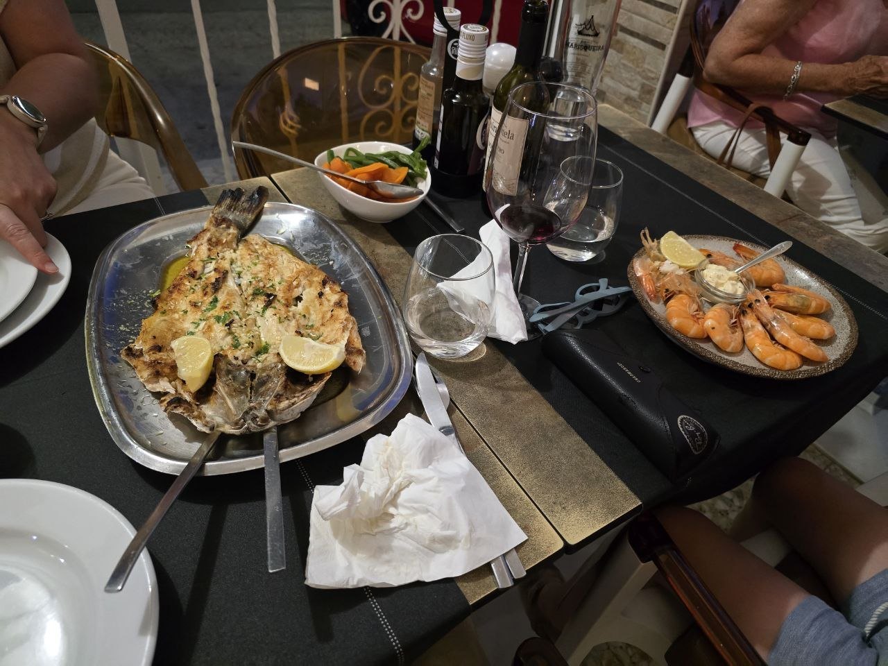 A restaurant table with grilled whole sea bass, boiled shrimp, vegetables, wine, water, and bread