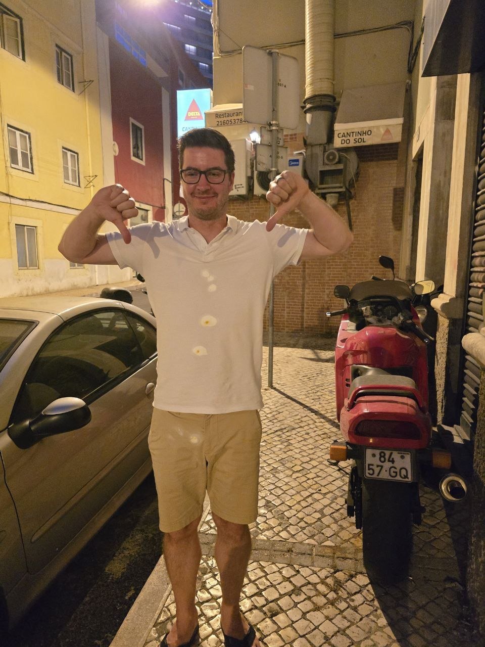 Ben standing on a Lisbon street giving two thumbs down with tomato sauce spots on his white polo after dinner