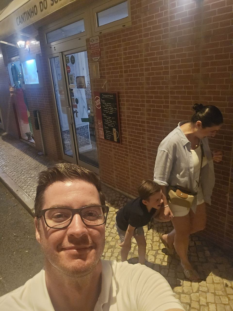 Ben taking a smiling selfie on the Lisbon sidewalk while Catherine and Noah laugh outside Cantinho do Sol