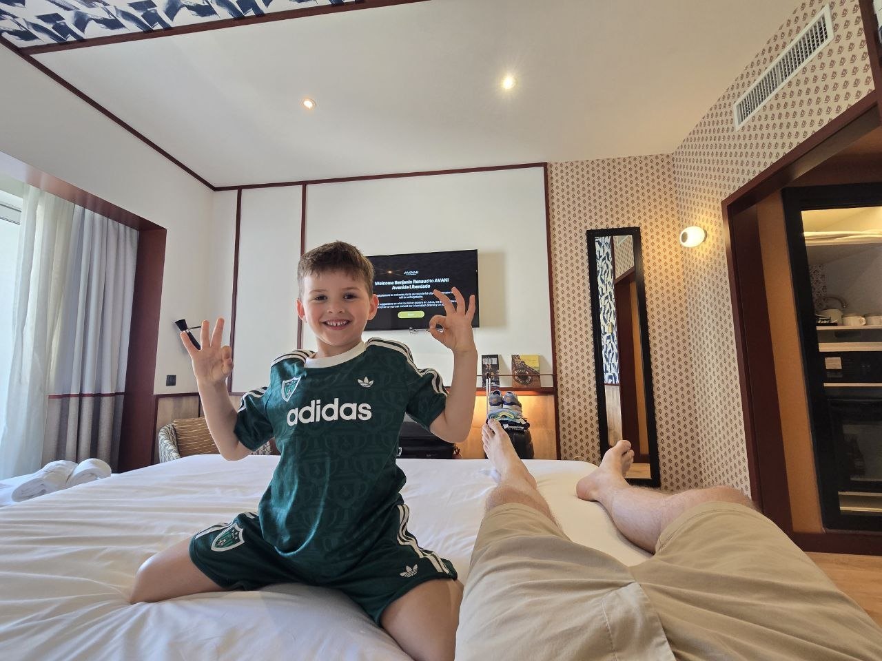 Noah smiling on the bed in the final Lisbon hotel room wearing his green kit