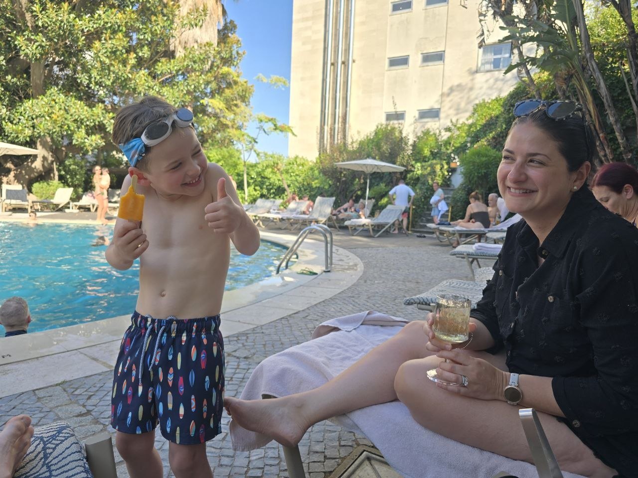 Noah giving a thumbs-up with a popsicle beside the Lisbon hotel pool while Catherine smiles from a lounger