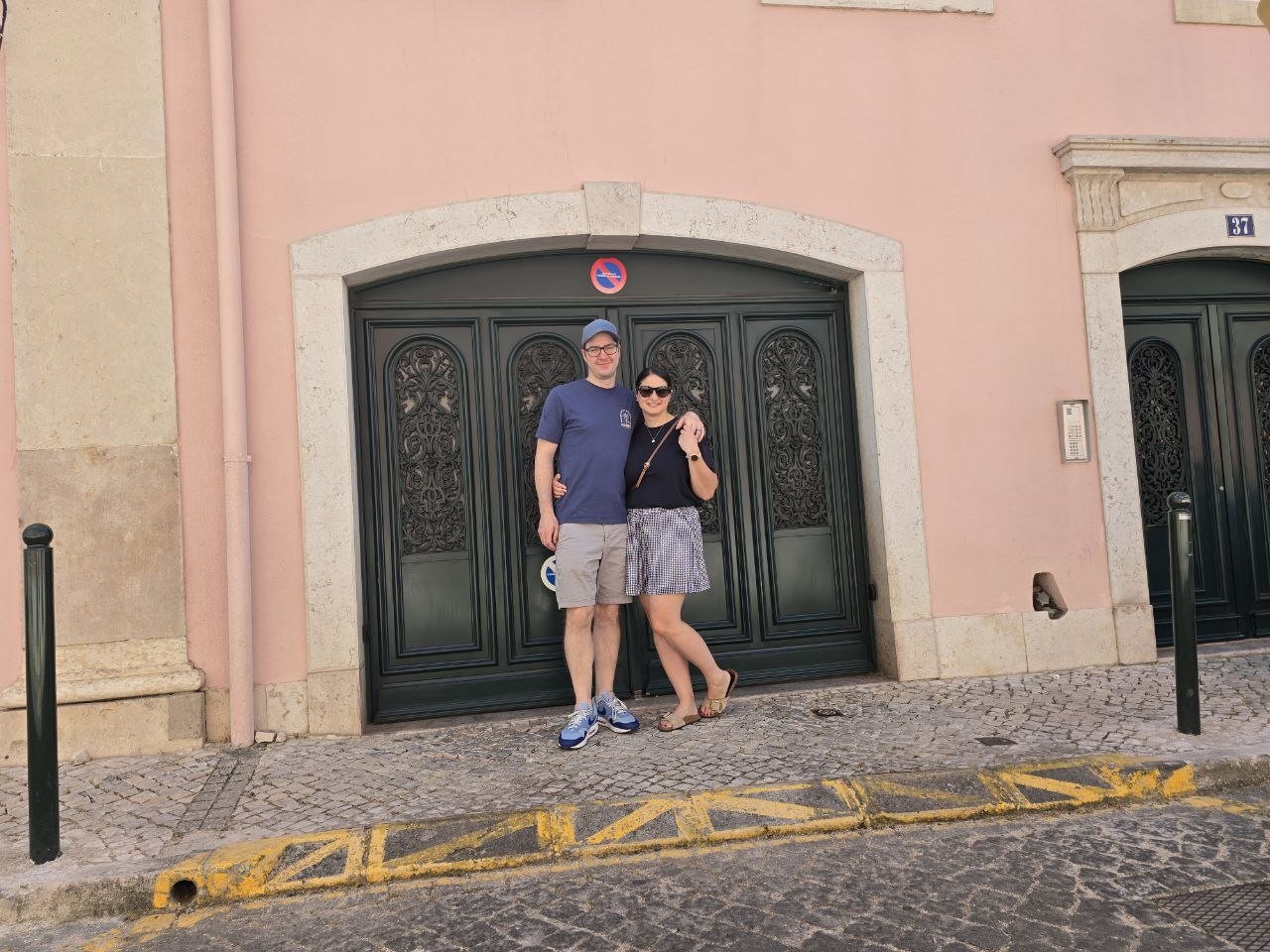 Benji and Catherine standing together in front of an ornate black door on a pink Lisbon street