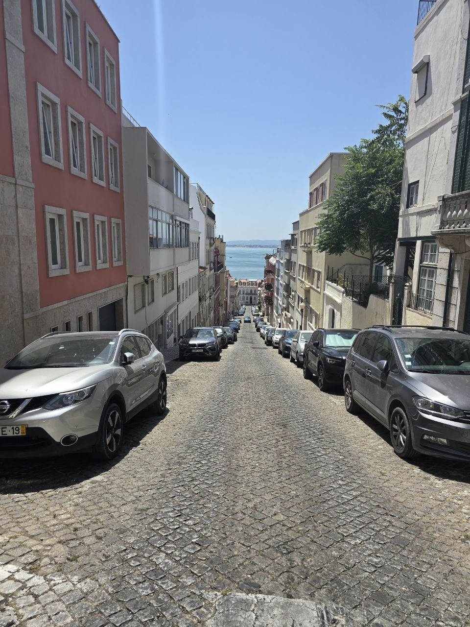 Steep cobblestone Lisbon street with parked cars and the Tagus River visible at the bottom