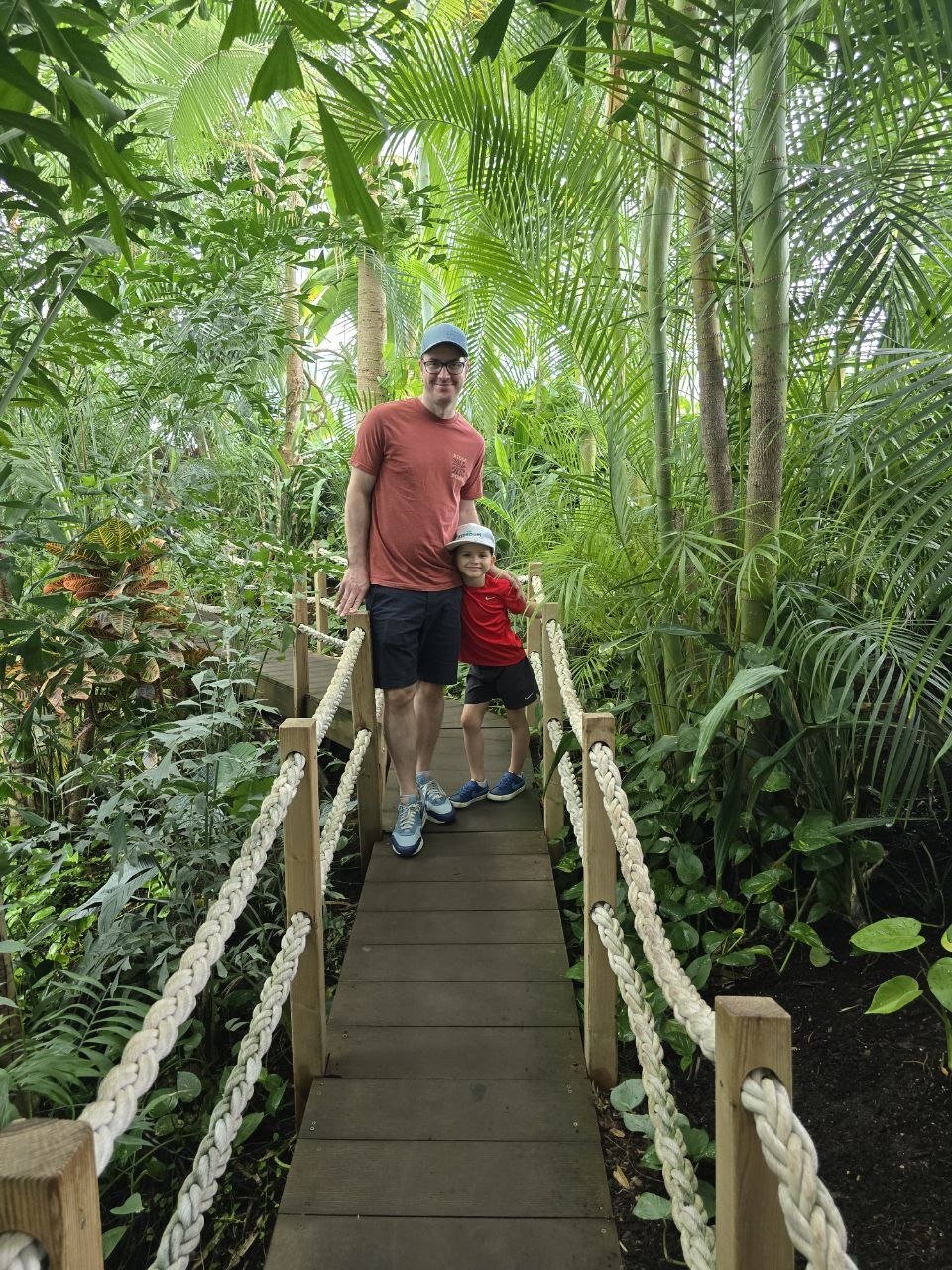 Benji and Noah standing together on a rope-lined wooden bridge surrounded by lush tropical plants at the Oceanário