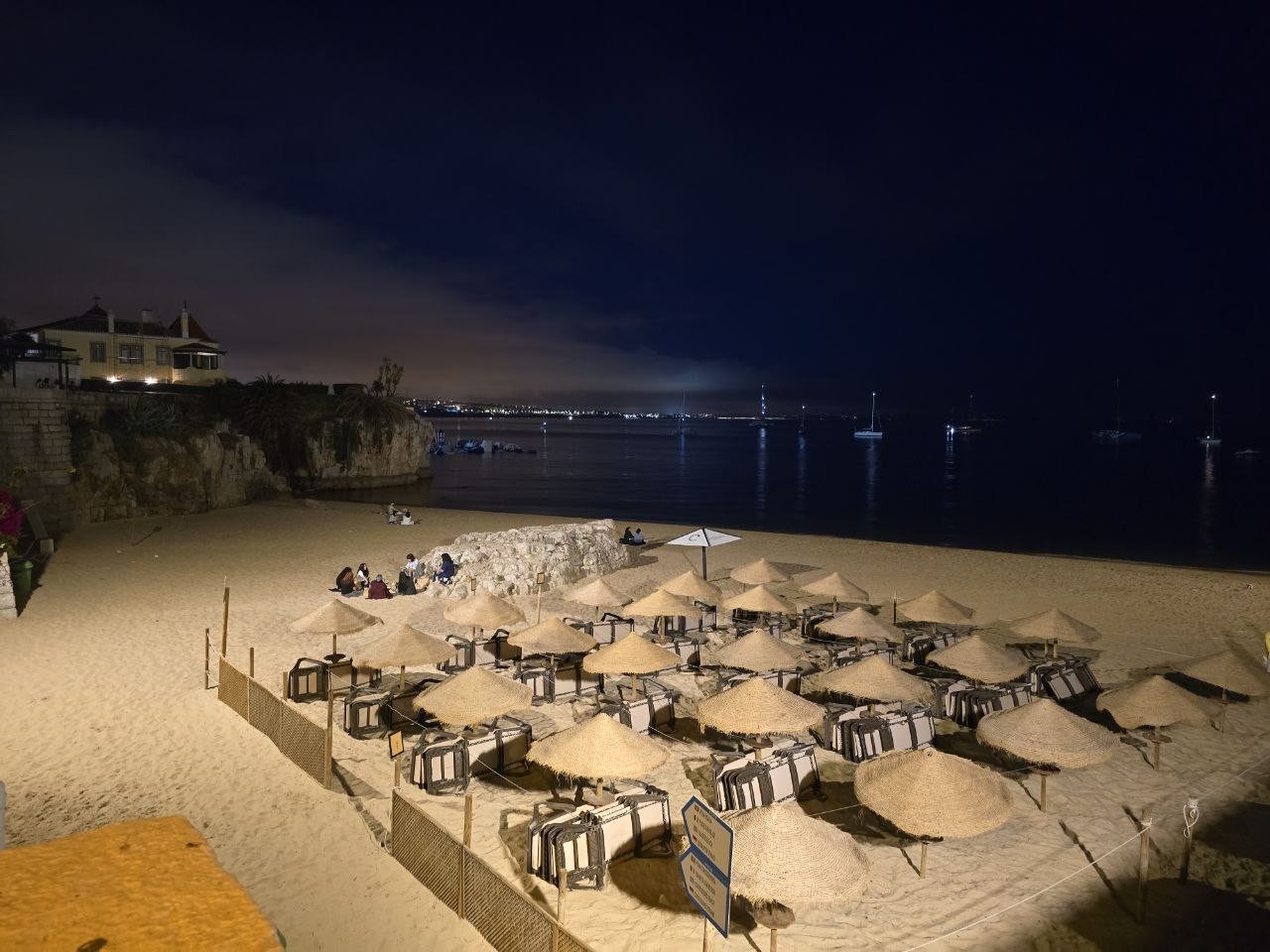 Cascais beach at night with straw umbrellas, boats, and lights on the water