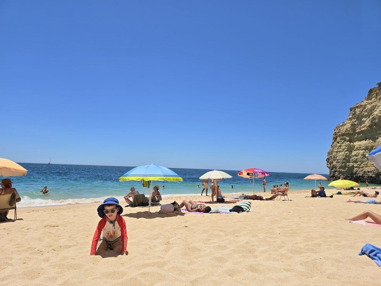 Noah playing in the sand at Vale de Centeanes Beach with umbrellas, cliffs, and blue ocean behind him